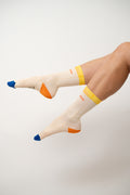 Cream El Guilty Pleasure socks with yellow cuffs and blue toes, legs raised against white